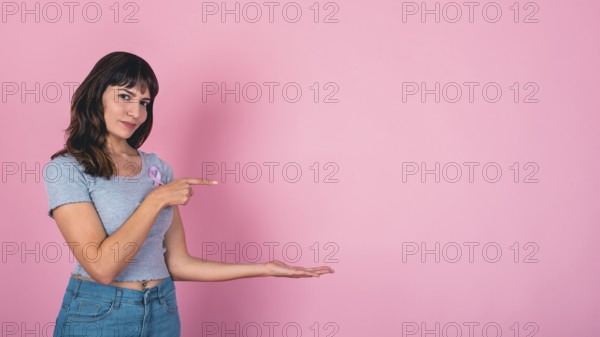 Young woman pointing and presenting with a pink ribbon for breast cancer awareness and copy space while looking at camera