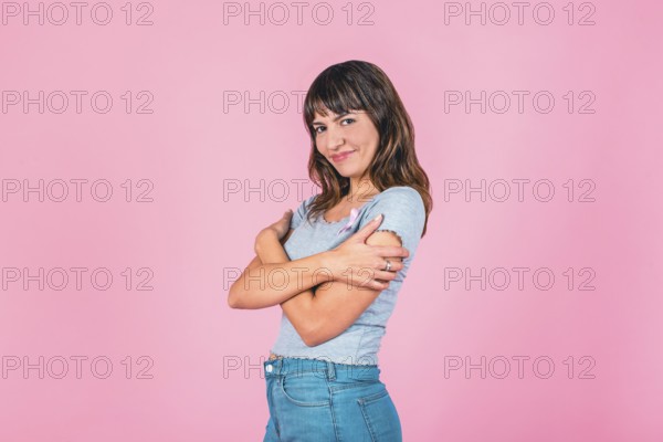 Young woman embracing herself with a pink breast cancer awareness ribbon on a pink backdrop