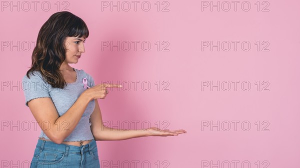 Young woman pointing and presenting with a pink ribbon for breast cancer awareness and copy space
