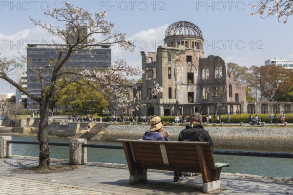 Tourists sitting on a bench under blossoming cherry trees, Sakura, Atomic Bomb Dome behind them, ruins of the former Chamber of Commerce and Industry building, landmark, UNESCO World Heritage Site, peace memorial and memorial to the victims of the atomic bombing of Hiroshima in the Peace Park on the Otagawa River, Naka Ward, Hiroshima, Japan