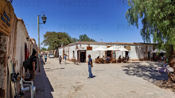 People sit under umbrellas on a lively street next to typical local buildings in the sunshine, The town of San Pedro de Atacama in Chile