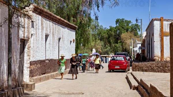 Pedestrians in summer clothes walk along a street with white buildings and a red car, The town of San Pedro de Atacama in Chile