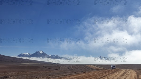 A dusty road leads through a remote desert landscape, bordered by mountains and clouds under a clear blue sky, The landscape at San Pedro de Atacama in Chile