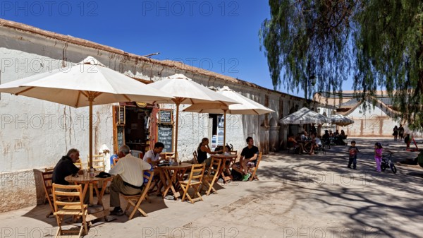 People sit in a café under parasols on the sunny street next to an old building, The town of San Pedro de Atacama in Chile