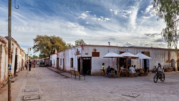 People move along a street with old buildings under partly cloudy skies and cyclists, The town of San Pedro de Atacama in Chile