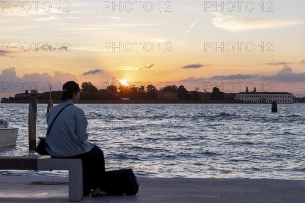 Venice, Italy - 2 September 2025: Passengers sit on the jetty at the Lido at sunset, waiting for the arrival of a boat to take them to another island, while a film poster is seen in the foreground, during the 82nd Venice International Film Festival