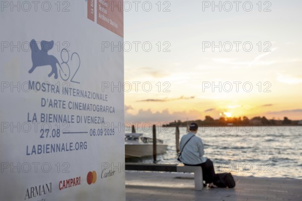 Venice, Italy - 2 September 2025: Passengers sit on the jetty at the Lido at sunset, waiting for the arrival of a boat to take them to another island, while a film poster is seen in the foreground, during the 82nd Venice International Film Festival