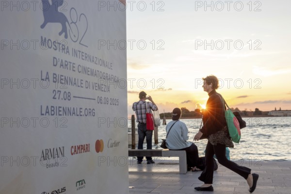 Venice, Italy - 2 September 2025: Passengers sit on the jetty at the Lido at sunset, waiting for the arrival of a boat to take them to another island, while a film poster is seen in the foreground, during the 82nd Venice International Film Festival