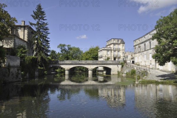 A serene scene with a stone bridge over a reflective river, surrounded by historic buildings and trees under a blue sky. The Seugne in Jonzac in Charente in France