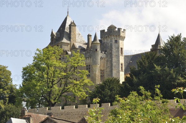 A medieval stone castle with towers surrounded by lush greenery under a clear sky. Village of Montfort in Périgord in France