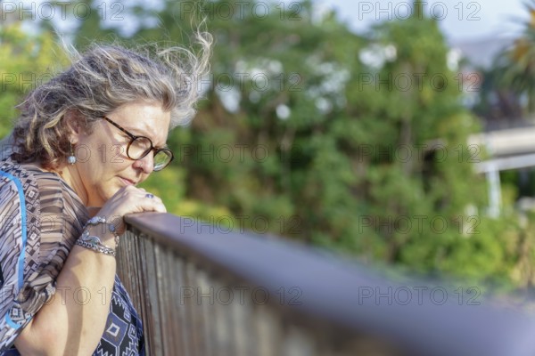 Grey-haired senior woman wearing glasses, leans on a railing, lost in contemplation amidst a serene natural backdrop