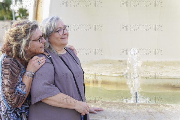 Two happy senior women hugging each other near a fountain in a city park, enjoying a sunny day together