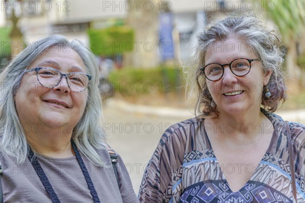 Two mature women with gray hair and glasses are smiling outdoors, possibly enjoying a leisurely day together