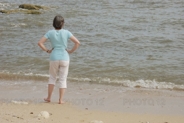 A woman stands on a sandy beach looking at the ocean waves, with rocks in the background in Royan in Charentes, France