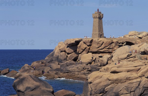 A lighthouse stands on a rocky coastline with people nearby and a clear blue sky. The granit rose coast in ploumanach in Brittany, France
