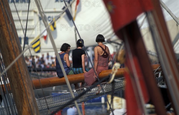 Three women sitting on a sailboat's wooden deck surrounded by nautical flags and ropes. Maritime festival in Brittany