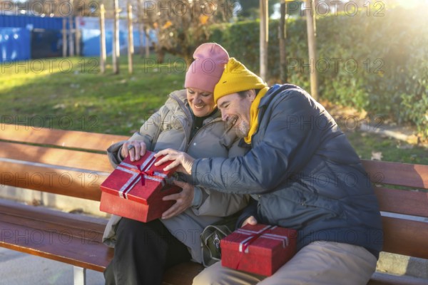 Elderly couple exchanging christmas gifts while sitting on a bench in a city park during a sunny winter day