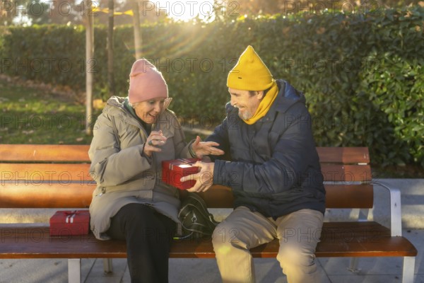 Elderly man surprising his wife with a festive christmas gift while sitting together on a park bench, enjoying a warm winter day