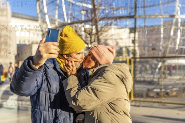 Happy senior couple kissing and taking a selfie with smartphone in a winter city during christmas holidays