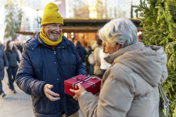Happy senior man presenting a christmas gift to his wife while enjoying the festive atmosphere of a bustling holiday market in the city