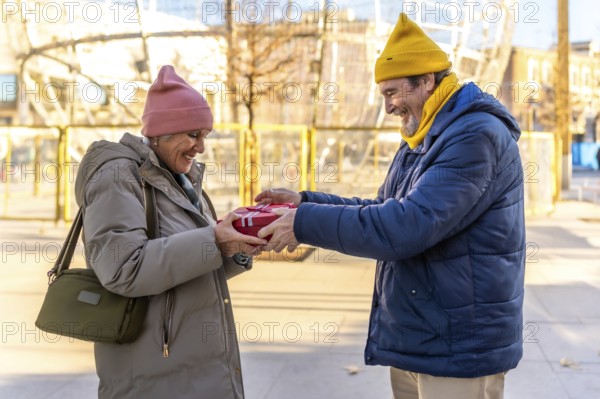 Happy senior man giving christmas present to smiling woman in amusement park during winter holidays