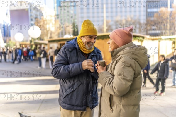 Smiling senior couple holding hot beverages and enjoying each other's company at a bustling christmas market in a winter city setting