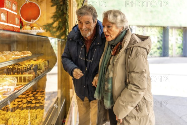 Happy retired couple selecting delicious sweets and cakes at a charming wooden stall in a festive christmas market during winter