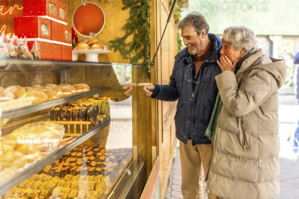 Joyful elderly couple pointing at delicious pastries in a bakery display window at an outdoor christmas market, enjoying festive holiday shopping