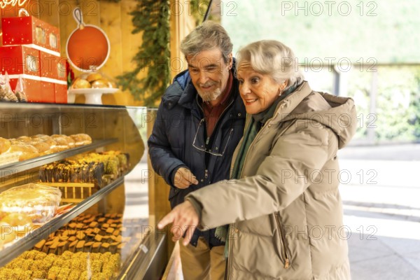 Happy senior couple pointing and choosing delicious pastries from a display at an outdoor christmas market