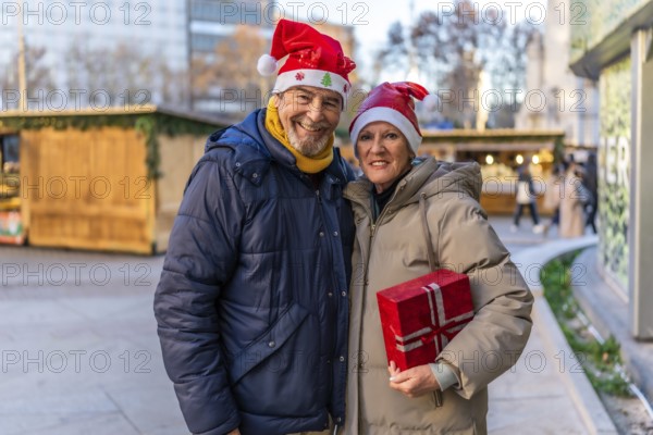 Smiling senior couple wearing santa hats holding christmas present at a christmas market in the city
