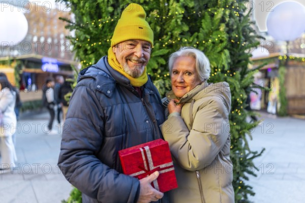 Smiling senior couple holding christmas gift, enjoying christmas market in the city during winter holidays
