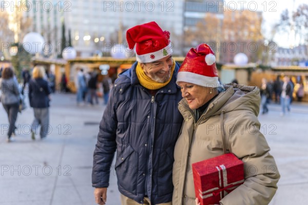 Smiling senior couple wearing santa hats holding hands and enjoying christmas market in the city during winter holidays