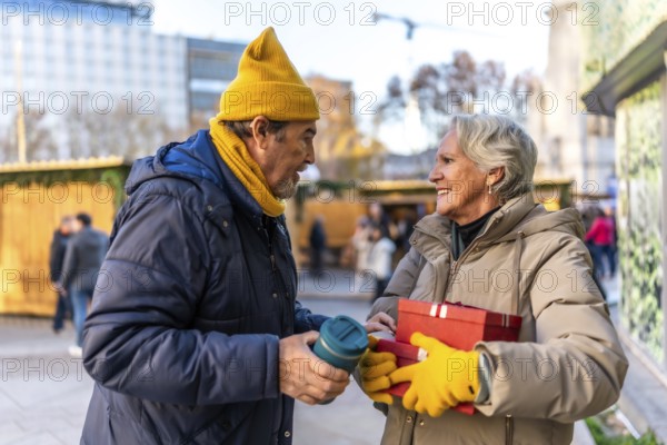 Happy senior couple exchanging christmas gifts and enjoying a warm beverage at a bustling outdoor christmas market in the city