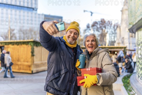 Elderly couple taking a selfie with smartphone at a christmas market, holding gifts and hot drinks, enjoying winter holidays in the city