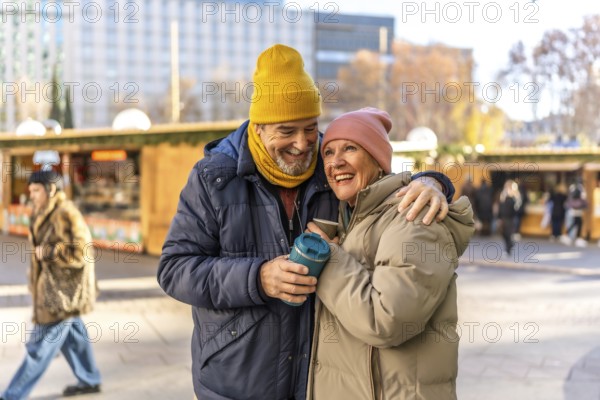 Elderly couple sharing warm drinks while enjoying the festive atmosphere of an outdoor christmas market, embracing and smiling joyfully