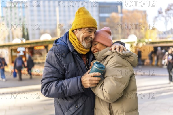 Elderly couple hugging and smiling at an outdoor christmas market, enjoying a warm beverage on a cold winter day