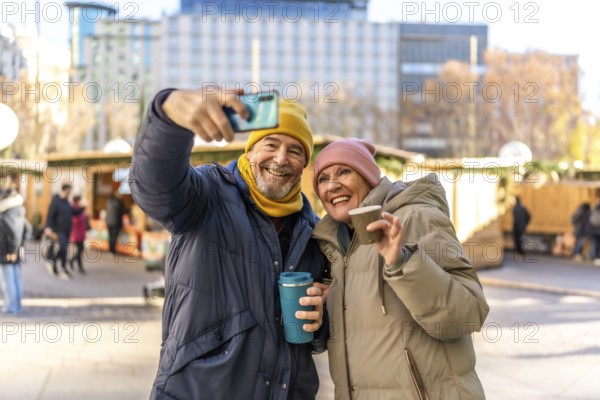 Smiling senior couple taking a selfie and drinking hot beverages at a christmas market in the city