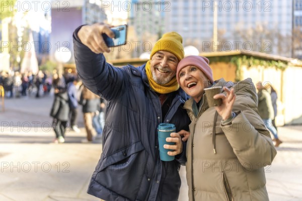 Smiling senior couple taking selfie with smartphone while enjoying hot drinks at a christmas market in winter city