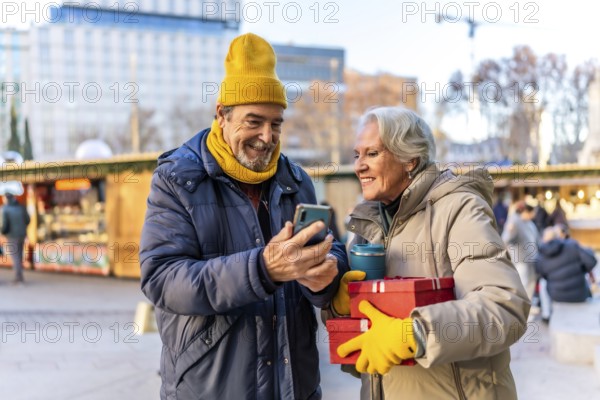 Happy retired couple looking at smartphone and holding christmas gifts and reusable coffee cup at a winter street market