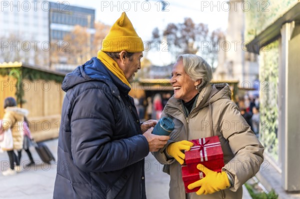Smiling elderly couple exchanging christmas presents and enjoying hot beverages at a winter market, celebrating the festive season together