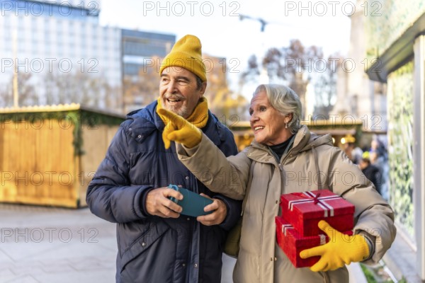 Cheerful senior couple holding christmas gifts and pointing at something interesting at the christmas market in the city