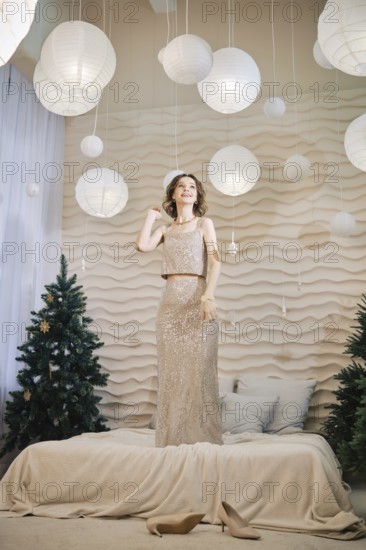 A woman poses gracefully in a shimmering dress on a bed covered with a soft blanket. She stands under hanging paper lanterns, with decorated trees in the background, creating a festive atmosphere