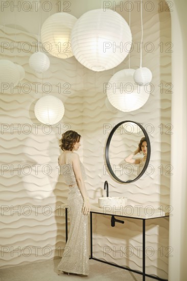 A woman dressed in a sparkling gown admires her reflection in a round mirror. Soft, textured walls and pendant lights create a serene atmosphere in a modern bathroom