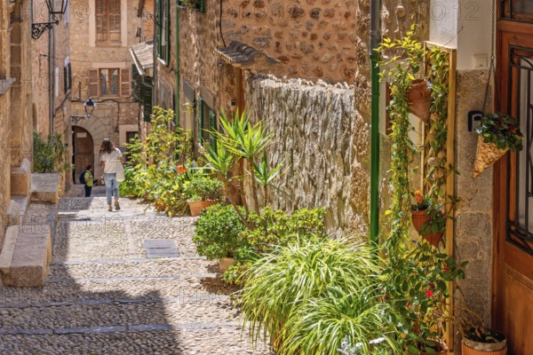 Alone woman walking in an idyllic cobblestone alley in an old village with green plants and flowers in pots outside a door on a sunny summer day, Fornlalutx, Mallorca, Spain