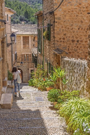 Woman walking in an idyllic cobblestone alley in an old village with green plants and flowers in pots, Fornlalutx, Mallorca, Spain