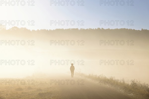 Alone person in silhouette walking on a dirt road in the countryside in the morning fog