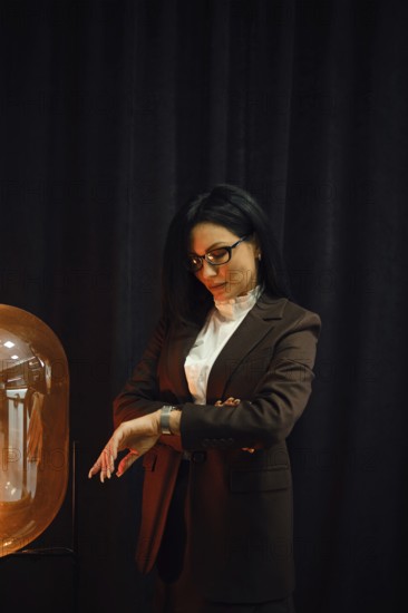 A woman dressed in a fashionable brown suit stands near a glowing lamp. She checks her watch with a contemplative expression, set against a dark backdrop that contrasts with the warm light