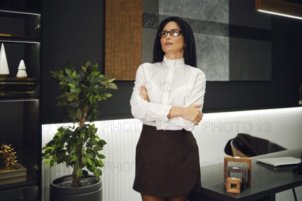 A confident woman stands with her arms crossed in a modern office. She wears a white blouse and a dark skirt, surrounded by greenery and contemporary decor