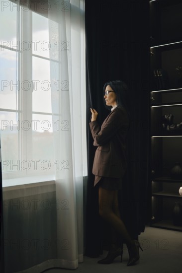 A woman dressed in a tailored suit stands by a large window, pulling back sheer curtains. She appears thoughtful as she gazes outside, surrounded by elegant, dark furnishings