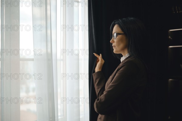 A businesswoman stands by a window in a modern office, gazing thoughtfully outside. She wears glasses and a stylish outfit, with soft natural light illuminating the room